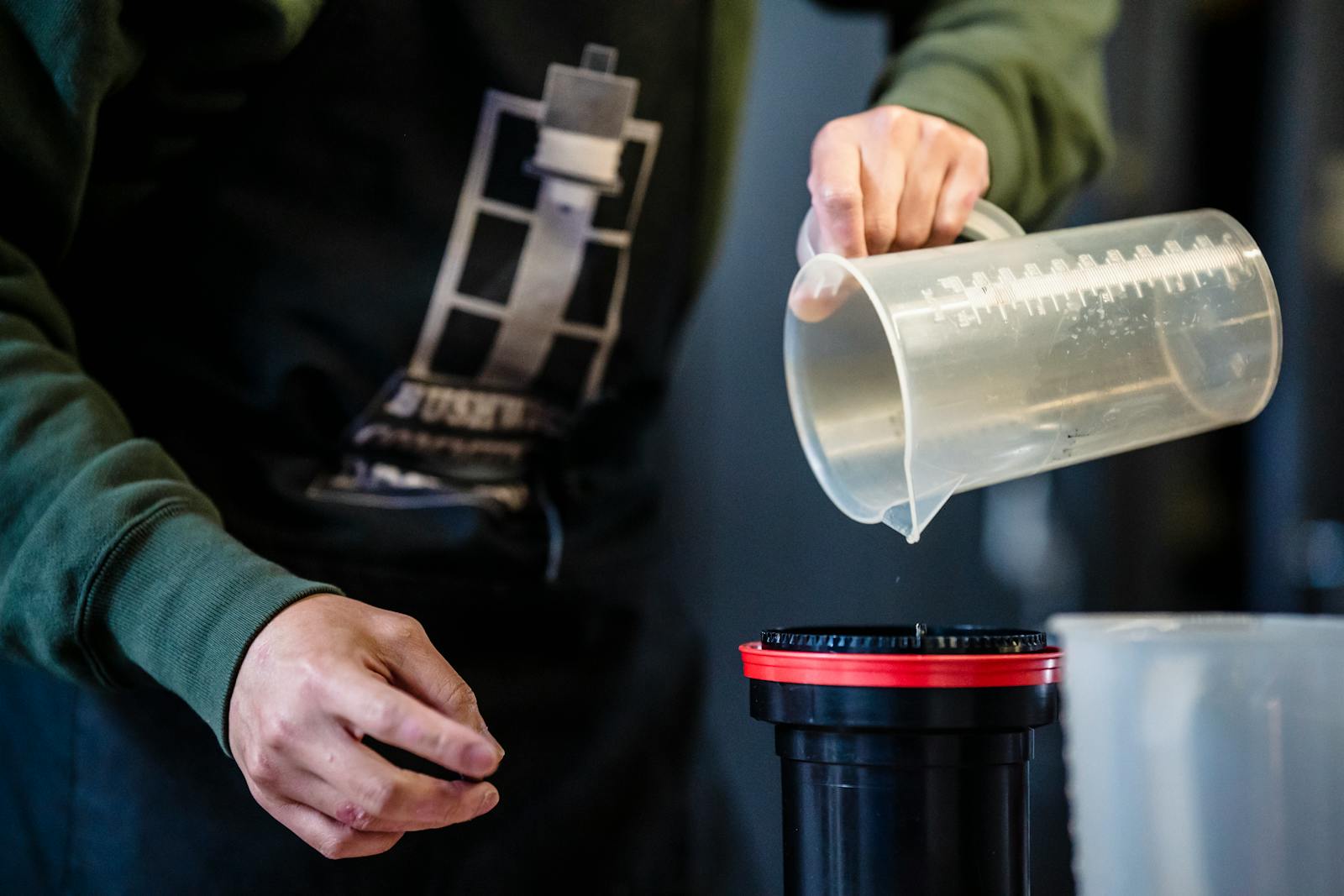 Hands pouring liquid from a measuring cup into a container, possibly for photo development.